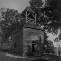 Black-and-white photograph of a small, square wooden watch house with a pitched roof and open bell tower on top, set among trees, with a door and small window visible—associated with Revolutionary-era signaling or lookout structures.