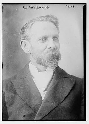 Black-and-white portrait photograph of a middle-aged man identified as Rev. Frank Sandford. He has short light-colored hair, a full beard and mustache, and wears a dark suit jacket with a high white collar. He is posed in three-quarter profile, looking slightly upward and away from the camera.
