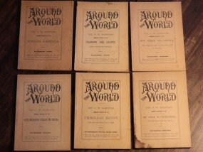 Six worn vintage booklets titled “Around the World” are arranged in two rows on a dark surface. The tan covers feature decorative black lettering and subtitles for different issues or topics. The booklets show signs of age, including creases, discoloration, and frayed edges.