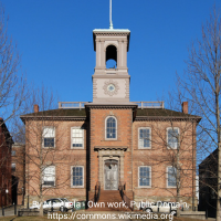 Historic brick building with a central clock tower and arched belfry, topped by a flagpole. The symmetrical façade features tall windows and a central doorway with steps. Leafless trees and neighboring buildings frame the scene under a clear blue sky.