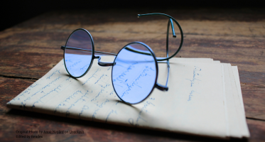Round, blue-tinted eyeglasses rest on a stack of handwritten papers on a wooden surface. The lenses reflect cursive writing, and the background is softly blurred, creating a quiet, scholarly atmosphere.