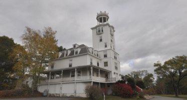 Large white historic building with wraparound porches and a tall octagonal tower rising above the roofline. The structure sits beside a curved paved road, surrounded by autumn trees and shrubs. Dark gray clouds fill the sky, giving the scene an overcast atmosphere.