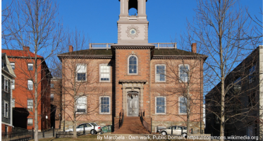 Historic brick building with a central clock tower and arched belfry, topped by a flagpole. The symmetrical façade features tall windows and a central doorway with steps. Leafless trees and neighboring buildings frame the scene under a clear blue sky.
