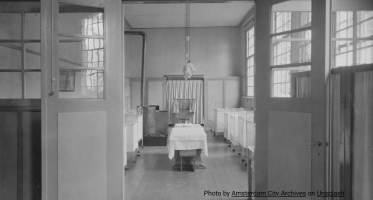 Black-and-white photograph of a hospital ward viewed through open double doors, with rows of metal bassinets or beds lining the room and a treatment table in the center.