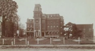Historic photograph of a large, multi-story brick institutional building with a central tower, identified as St. Louis’s Social Evil Hospital. A fence and open grounds surround the building, with trees visible on either side.