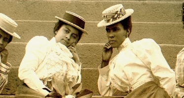 Sepia-toned photograph of four young women posing on outdoor stone steps. They wear long skirts, high-collared blouses with puffed sleeves, and decorative hats. Each leans on one arm with a composed expression, creating a staged, fashion-focused portrait from the late 19th or early 20th century.